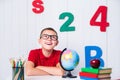 Happy cute clever boy is sitting at a desk in a glasses with raising hand. Child is ready to answer with a blackboard on Royalty Free Stock Photo