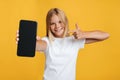 Happy cute european teen blonde girl shows finger at smartphone with blank screen