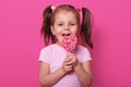 Happy cute girl wears rose t hirt, stands isolated over pink background, holds bright lollipop in hands. Cheery child with opened