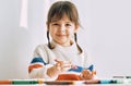 Happy cute little girl paints with oil pencils, sitting at white desk at home. Pretty smiling preschool kid draws Royalty Free Stock Photo