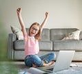 Happy cute little girl 8 years old with glasses sits at home on a carpet in front of a laptop, hands up Royalty Free Stock Photo