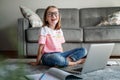 Happy cute little girl 8 years old in a striped t-shirt and jeans with glasses sits at home on a carpet in front of a laptop, Royalty Free Stock Photo