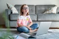 Happy cute little girl 8 years old in a striped t-shirt and jeans with glasses sits at home on a carpet in front of a laptop, Royalty Free Stock Photo