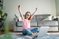 Happy cute little girl 8 years old in a striped t-shirt and jeans with glasses sits at home on a carpet in front of a laptop,