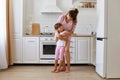 Happy cute small kid child daughter standing with mom having fun at home, standing in light kitchen room and hugging each other,