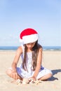 Happy cute teenage girl in a red Santa hat sitting on a sand on a sea beach and making a date 2020 from shells, vertical