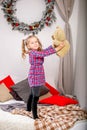 Happy cute young girl in a checkered blue-red dress standing on the bed with a teddy bear and holding it against the background of Royalty Free Stock Photo