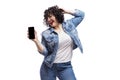 Happy dancing young beautiful girl in jeans shows a smartphone that is emitted on a black screen. Bright brunette with curly hair Royalty Free Stock Photo