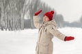 Happy dancing young woman enjoys nature in winter park. Young woman in down jacket and woolen red hat with mittens walks in snow