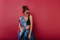 Happy dark-haired woman wears denim clothes spending time on photoshoot. Refined girl in sunglasses holding blue jacket