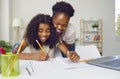 Happy dark-skinned mother watching her cheerful teenage daughter write and draw at home.