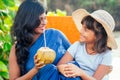 Happy daughter in straw hat with her beautiful mother in blue saree drinking fresh coconut water in tropical paradise