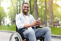 Happy disabled black man in wheelchair using smartphone, checking messages online at park