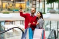 Happy diverse couple with gift bags using moving stairway, going shopping together at city mall Royalty Free Stock Photo