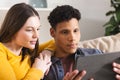 Happy diverse couple sitting on couch and using tablet in living room at home, copy space Royalty Free Stock Photo