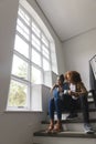Happy diverse couple sitting on stairs drinking coffee, embracing, looking out of window, copy space Royalty Free Stock Photo