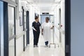 Happy diverse female doctor talking with senior female patient with drip in hospital corridor Royalty Free Stock Photo