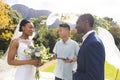 Happy diverse male officiant with bride and groom at outdoor wedding ceremony in sunny garden Royalty Free Stock Photo