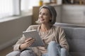 Happy dreamy mature woman holding tablet, sitting on couch
