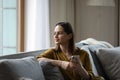 Happy dreamy millennial girl holding smartphone, resting on comfortable sofa