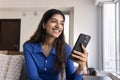 Happy dreamy young 20s Indian girl using smartphone on couch Royalty Free Stock Photo