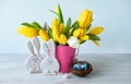 Happy Easter. Decorative two funny easter bunnies, eggs in bird nest and bouquet of yellow tulips in bucket on wooden table. Royalty Free Stock Photo