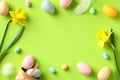 Frame made of colorful Easter eggs, daffodil spring flowers on green background. Flat lay,