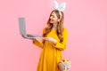 Happy Easter. woman in the ears of an Easter Bunny holding a basket of Easter eggs using a laptop on an pink background