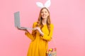 Happy Easter. Happy woman in the ears of an Easter Bunny holding a basket of Easter eggs using a laptop on an pink background