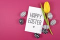 `Happy Easter` text card surrounded by black and white easter eggs and single yellow tulip spring flower on bright pink background Royalty Free Stock Photo