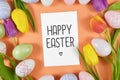 `Happy Easter` text card surrounded by colorful easter eggs and tulip spring flowers on orange background, flat lay Royalty Free Stock Photo
