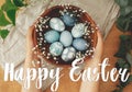 Happy Easter text. Easter greetings lettering. Hands holding wooden bowl with modern blue Easter eggs in spring flowers. Greeting Royalty Free Stock Photo