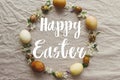 Happy Easter text on Easter eggs and spring flowers wreath on rustic linen, flat lay, handwritten sign. Beautiful stylish greeting Royalty Free Stock Photo