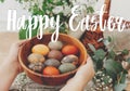 Happy Easter text. Easter greetings lettering. Hands holding wooden bowl with modern Easter eggs on rustic table with spring Royalty Free Stock Photo