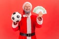 Happy elderly man with gray beard wearing santa claus costume looking at camera with excitement, holding soccer ball and money, Royalty Free Stock Photo