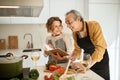 Happy elderly spouses checking recipe in cookbook while preparing healthy food together in kitchen, free space Royalty Free Stock Photo