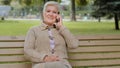 Happy elderly woman with beautiful face sitting on park bench takes phone call, gray-haired lady of retirement age