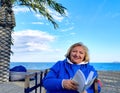 Happy elderly woman in blue clothes, sitting in a chair on the beach, looking at the camera with a background of sea and Royalty Free Stock Photo