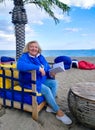 Happy elderly woman in blue clothes, sitting in a chair on the beach, looking at the camera, hold a book with a background of sea Royalty Free Stock Photo