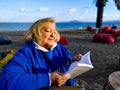 Happy elderly woman in blue clothes, sitting in a chair on the beach, reading a book against a background of sea and Royalty Free Stock Photo