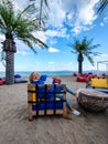 Happy elderly woman in blue clothes, sitting in a chair on the beach, reading a book with background of sea and palm Royalty Free Stock Photo