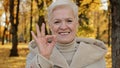 Happy elderly woman joyfully smiling standing in autumn park mature grandmother looking at camera showing gesture okay Royalty Free Stock Photo