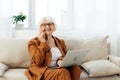 a happy elderly woman smiles sweetly sitting on the sofa in a bright apartment near the window and talking on a Royalty Free Stock Photo