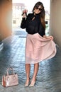 Happy elegant brunette woman with sunglasses wearing a pink pleated skirt ,black blouse, high pink-black heels, leather bag.