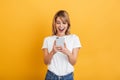 Happy emotional young blonde woman posing isolated over yellow wall background dressed in white casual t-shirt using mobile phone Royalty Free Stock Photo