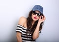 Happy enjoyment grimacing young woman in sunglasses and blue baseball cap posing and looking in striped blouse on blue empty Royalty Free Stock Photo