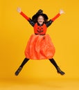 Happy ethnic girl in witch pumpkin costume jumping and celebrates Halloween on yellow background