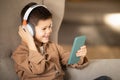 Happy european little boy in casual and wireless headphones watch video on smartphone, sits in armchair