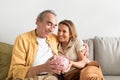 Happy european senior couple holding piggybank and embracing, sitting on sofa in living room interior, free space Royalty Free Stock Photo