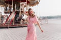 Happy european woman in long dress dancing near carousel. Outdoor shot of enchanting blonde girl in